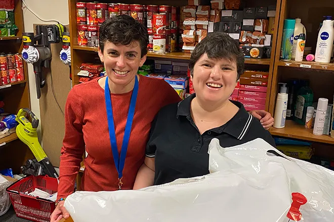 Two women standing together in a stocked pantry storeroom, one in a lanyard and one holding a large bag of donated goods, both laughing.