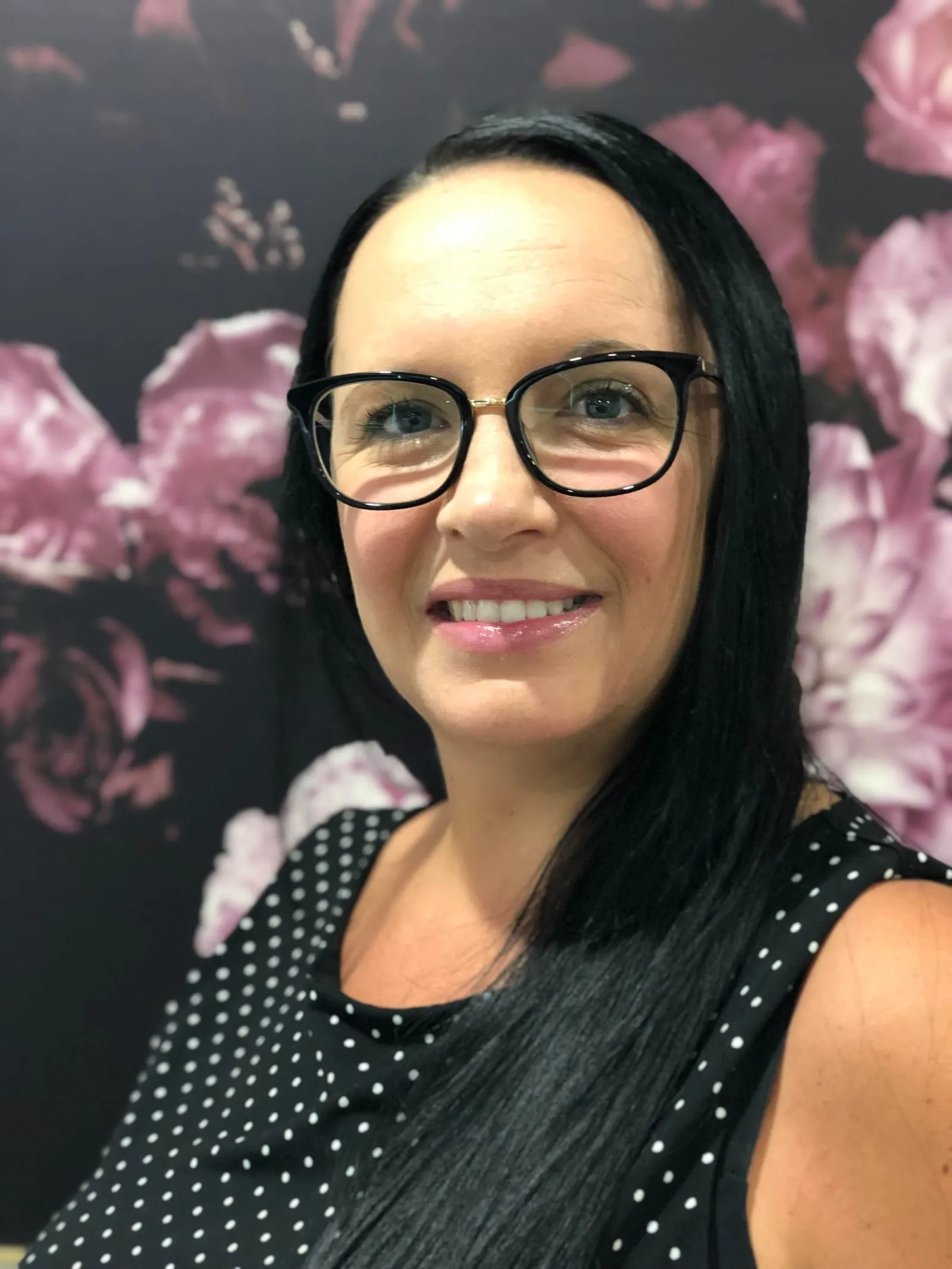 A woman in glasses and a black polka dot top smiling at the camera, in front of a dark floral wall.