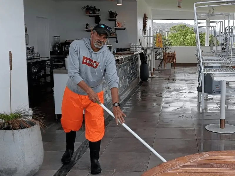 A man in orange waterproof overalls and gumboots hosing down the tiled floor of an outdoor cafe terrace, smiling at the camera.