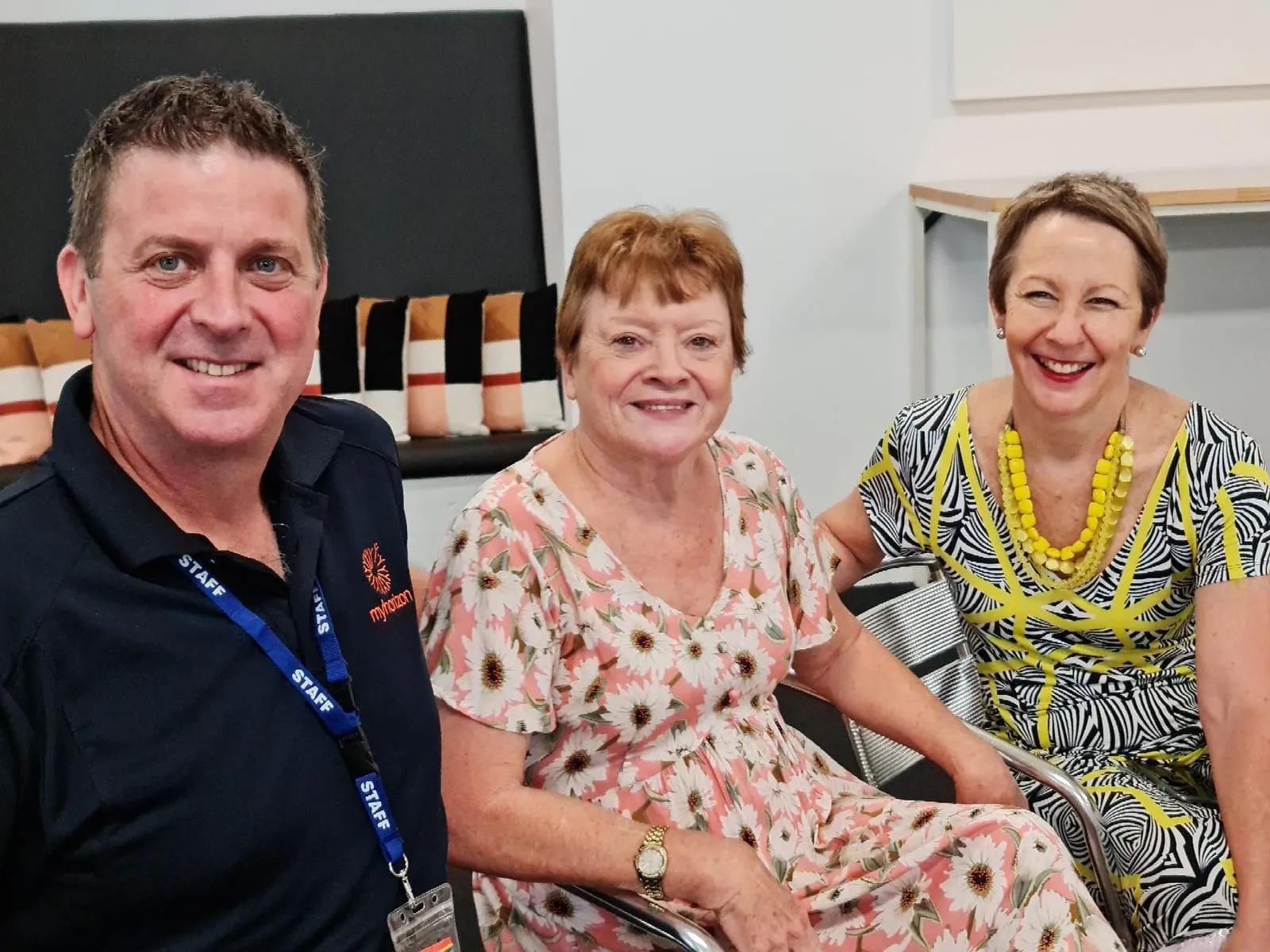 A man in a navy Myhorizon polo shirt and staff lanyard sitting with two women at a morning tea, all three smiling at the camera.
