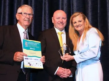 Two men and a woman on an awards stage, holding a glass trophy and a winner's certificate for The Bush Pantry.