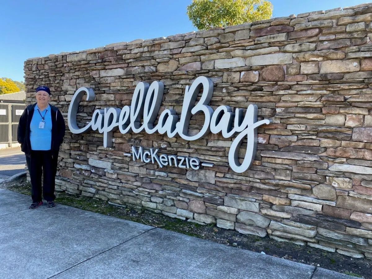 A woman in a cap and lanyard standing beside the stone entrance wall of Capella Bay McKenzie, on a clear day.
