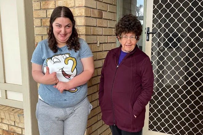 Two women standing together at the front door of a brick home, one giving a thumbs up.