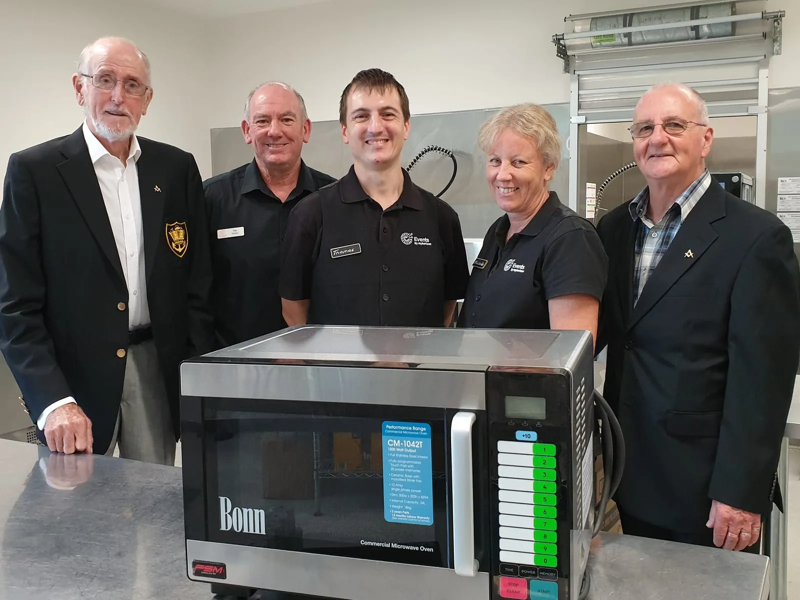 Five people standing behind a new commercial microwave oven in the Events by Myhorizon kitchen, three of them in kitchen uniforms and two in Freemasons blazers.