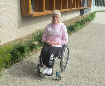 A young woman in a pink hoodie sitting in her wheelchair on a path outside a building, smiling at the camera.