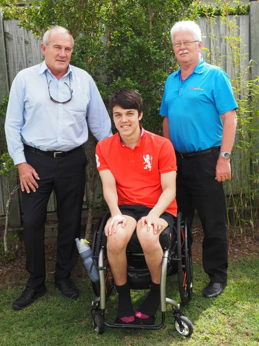 A young man in a red polo shirt sitting in his wheelchair on a lawn, with two men standing on either side of him.