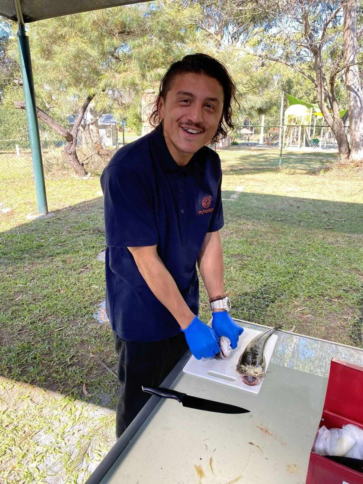 A young man in a navy Myhorizon polo shirt and blue gloves filleting a fish on a board at an outdoor bench, smiling at the camera.