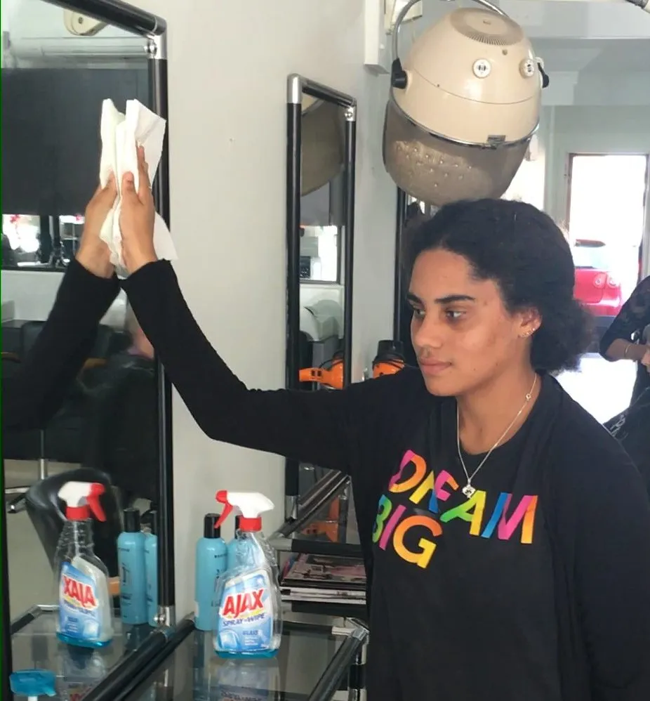 A young woman in a Dream Big t-shirt reaching up to polish a mirror in a hair salon, with cleaning products lined up on the glass shelf in front of her.