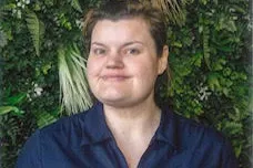 A woman in a navy work polo shirt smiling at the camera, in front of a green foliage wall.