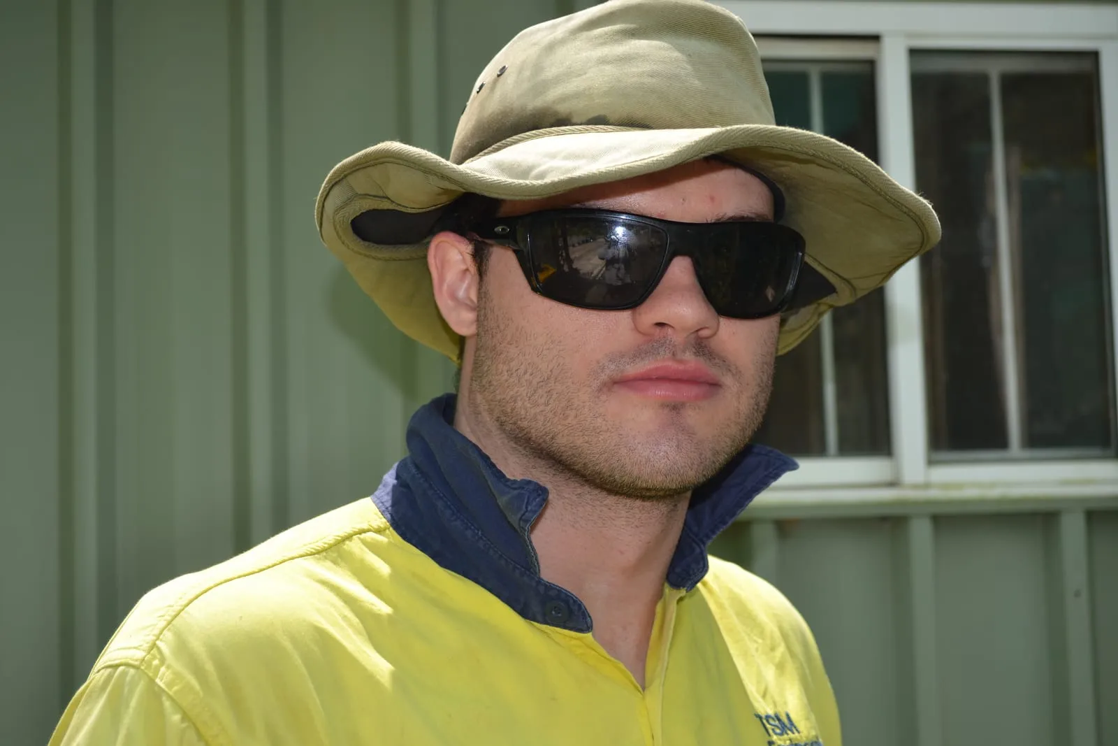 A young man in a wide-brimmed sun hat, sunglasses and a yellow high-visibility work shirt, outside a shed on a bright day.