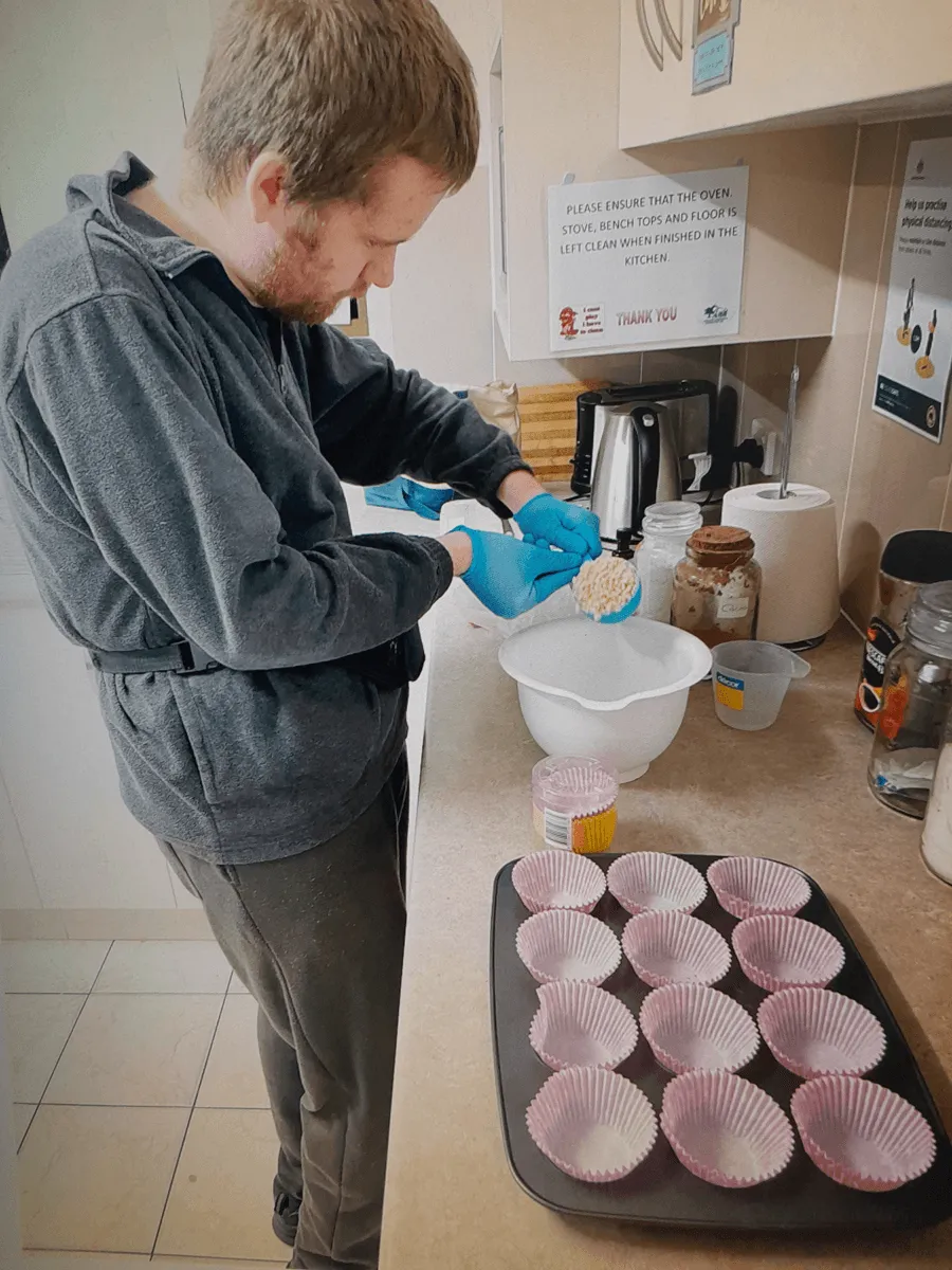 A young man in gloves spooning mixture into a bowl in a kitchen, with a tray of empty patty cases lined up on the bench in front of him.