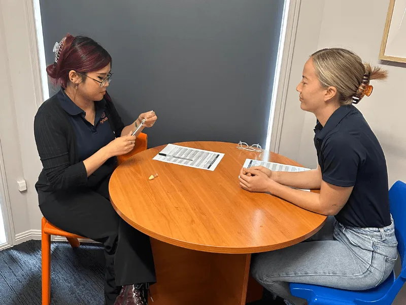 Two Myhorizon staff sitting across a round table in a training session, one demonstrating with a small device while the other watches, with printed handouts between them.