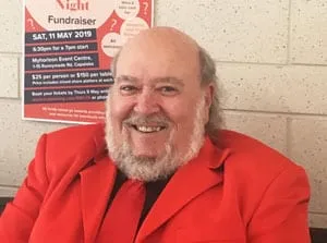 A man in a bright red jacket smiling at the camera, in front of a poster advertising a trivia night fundraiser at the Myhorizon Event Centre on Saturday 11 May 2019.