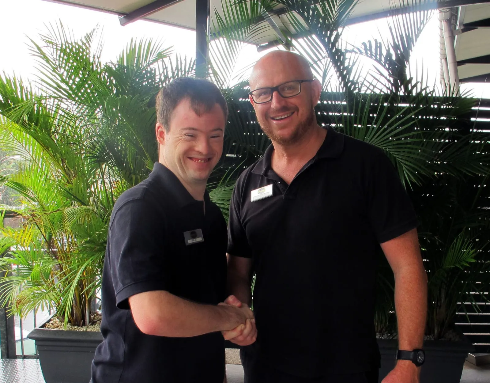 Two men in black work polo shirts shaking hands and smiling at the camera, standing in front of palms outside a workplace.