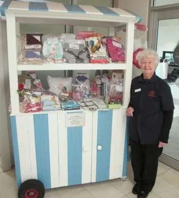 A woman in a Myhorizon uniform standing beside a blue and white striped market cart stacked with handmade quilts, blankets and craft goods.