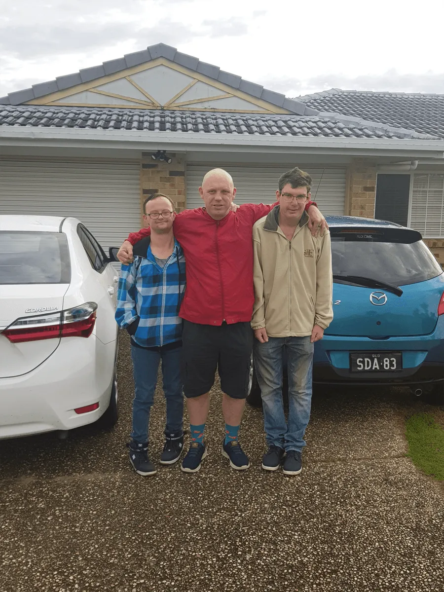 Three housemates standing arm in arm on the driveway in front of their home, with two cars parked either side of them.