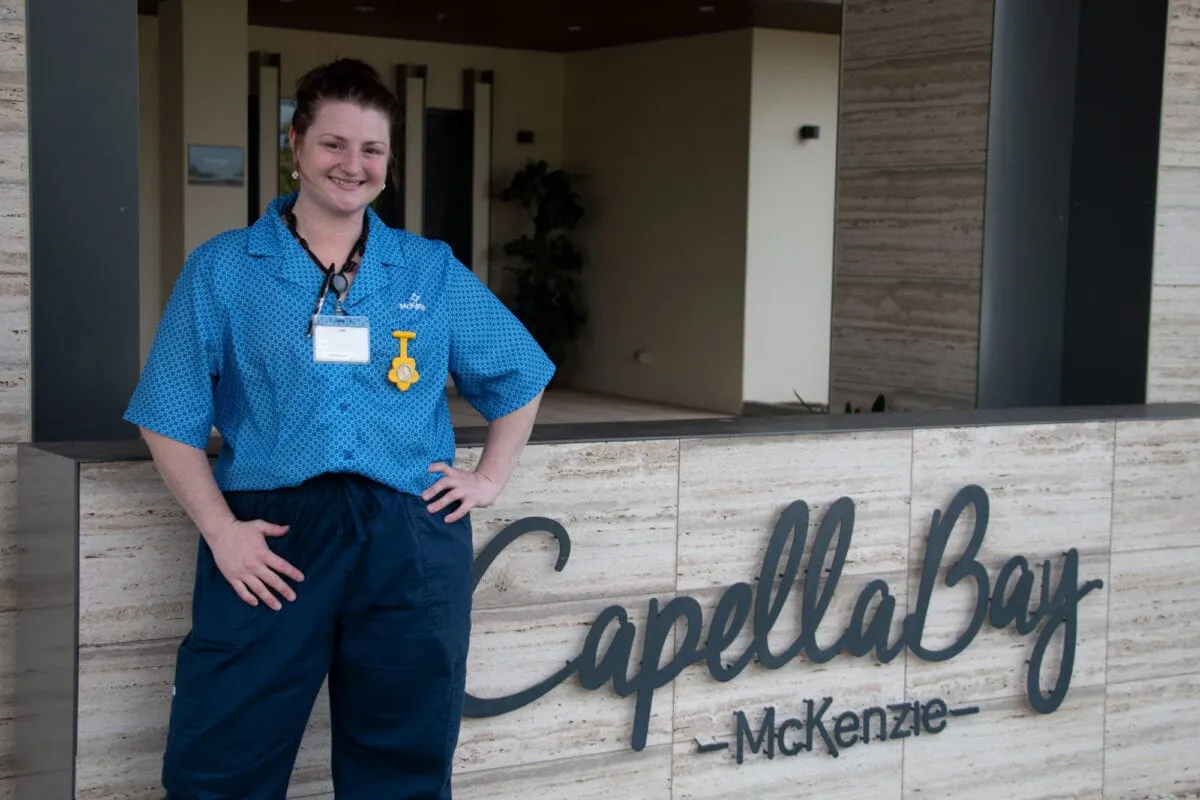 A woman in a blue care uniform with a fob watch and lanyard standing beside the Capella Bay McKenzie sign at the entrance to the building.