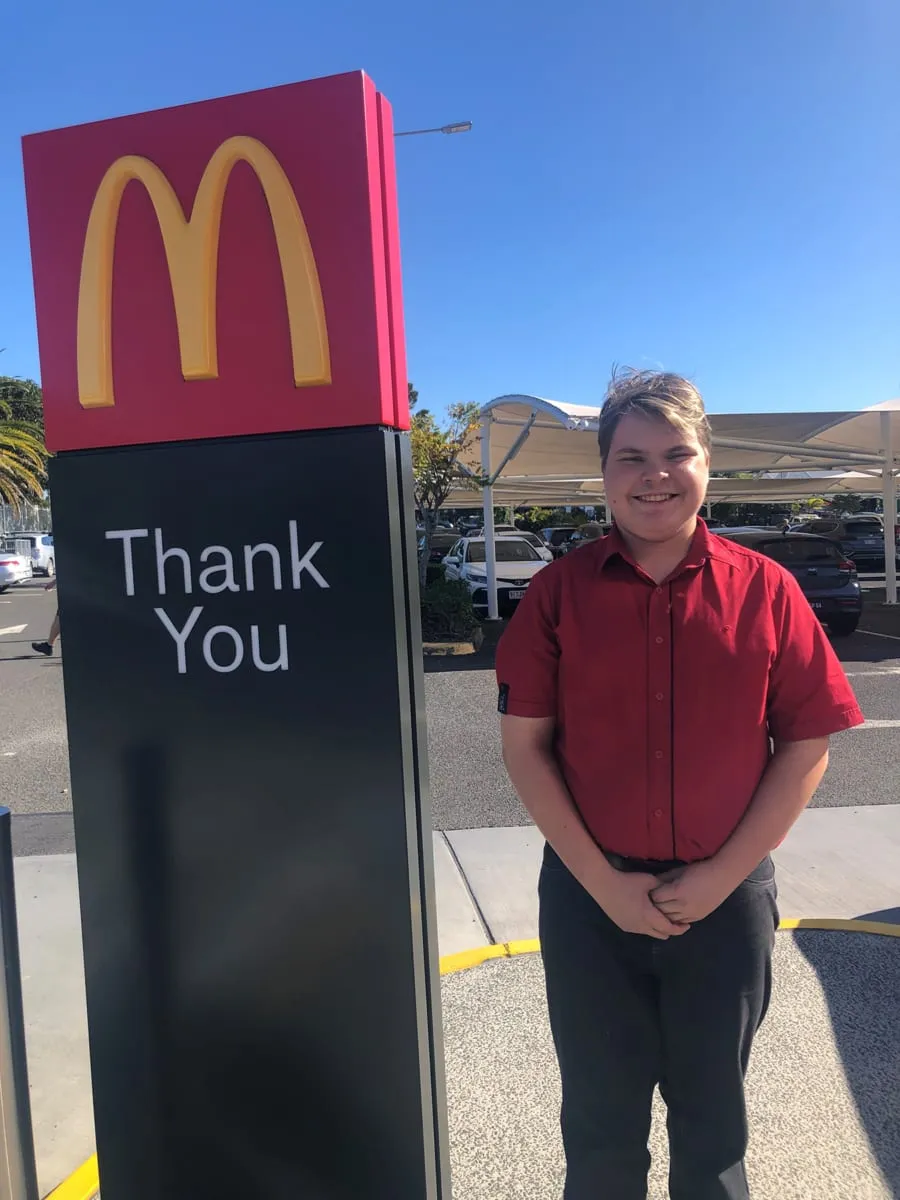 A young man in a red work shirt standing beside the drive-through Thank You sign at McDonald's, smiling in the sunshine.