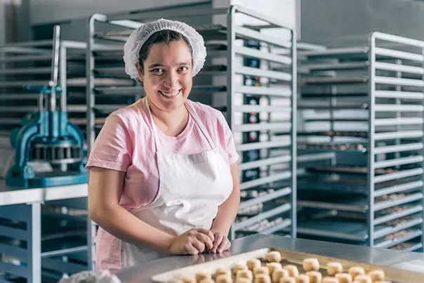 A woman in a hairnet and apron smiling at the camera in a commercial bakery, with a tray of pastries in front of her and cooling racks behind.