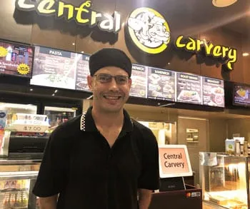A man in a black uniform and cap smiling behind the counter at Central Carvery, with menu boards lit up above him.