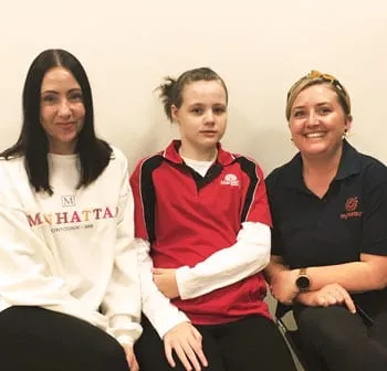 A girl in a red and black sports jacket sitting between two women, all three smiling at the camera.