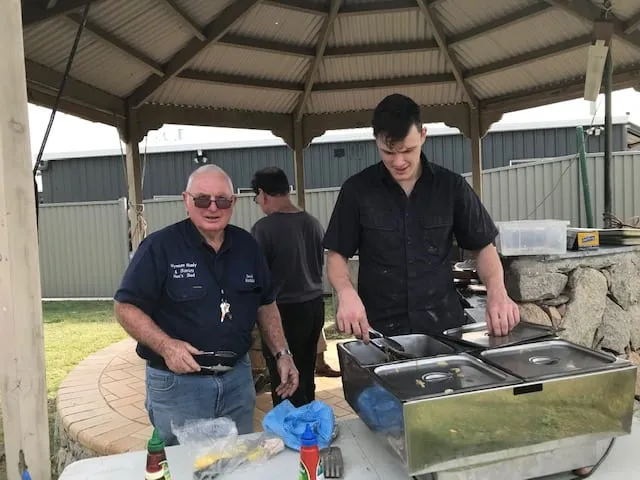 Two men cooking at a barbecue under a rotunda, one turning food on the hotplate while the other holds a pan.