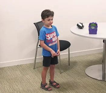 A young boy in a bright blue superhero t-shirt standing in a therapy room, with a chair and a small round table behind him.