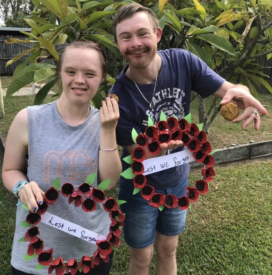 A young woman and a young man standing in a backyard, each holding a large ANZAC wreath they have made from egg cartons painted as red poppies, with Lest We Forget written across the middle.