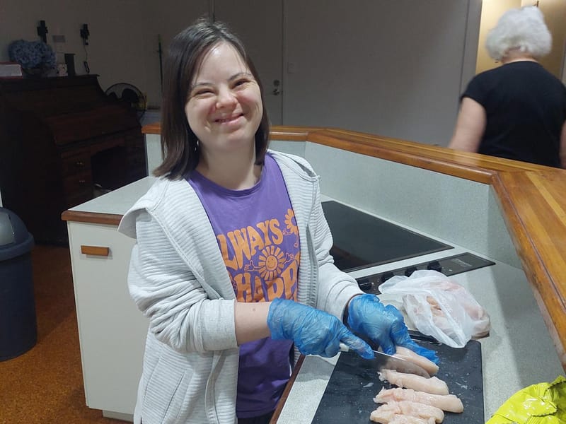A young woman in blue gloves smiling at the camera as she prepares chicken on a chopping board.