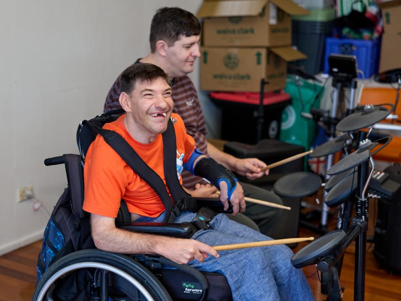 Two men playing electronic drums together, one laughing in his wheelchair.