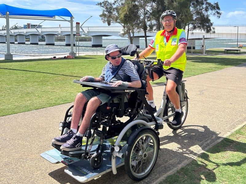 A man in his wheelchair riding on the front platform of an adaptive bike along a waterfront path, with a rider in a hi-vis vest pedalling behind.