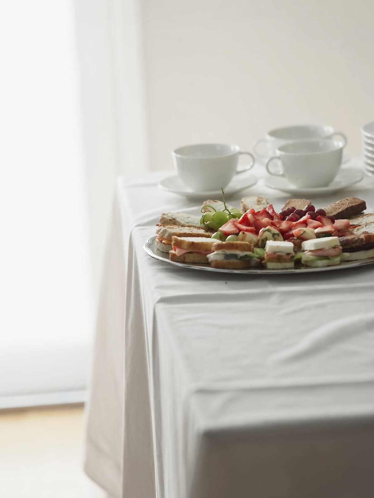 A platter of small sandwiches, strawberries and grapes on a white tablecloth, with teacups and saucers behind it