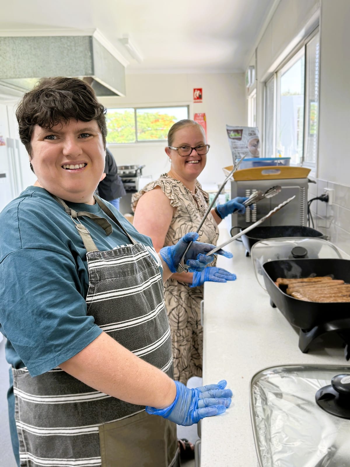 Two people in blue gloves cooking at a kitchen bench lined with electric frying pans, both smiling at the camera.