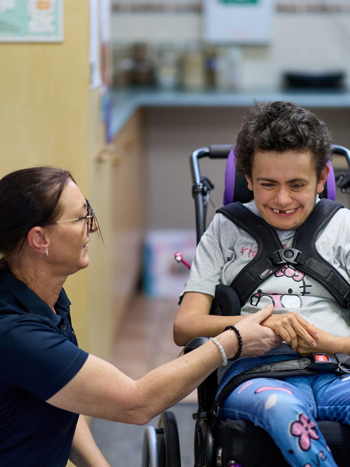 A support worker crouching beside a smiling young person in a wheelchair, the two of them holding hands.