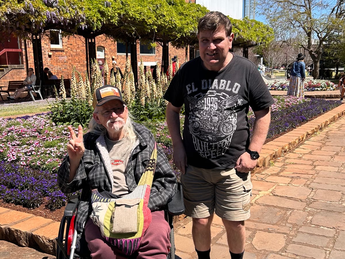 Two men at a flower festival, one giving a peace sign from his wheelchair, beside beds of pink and purple flowers.