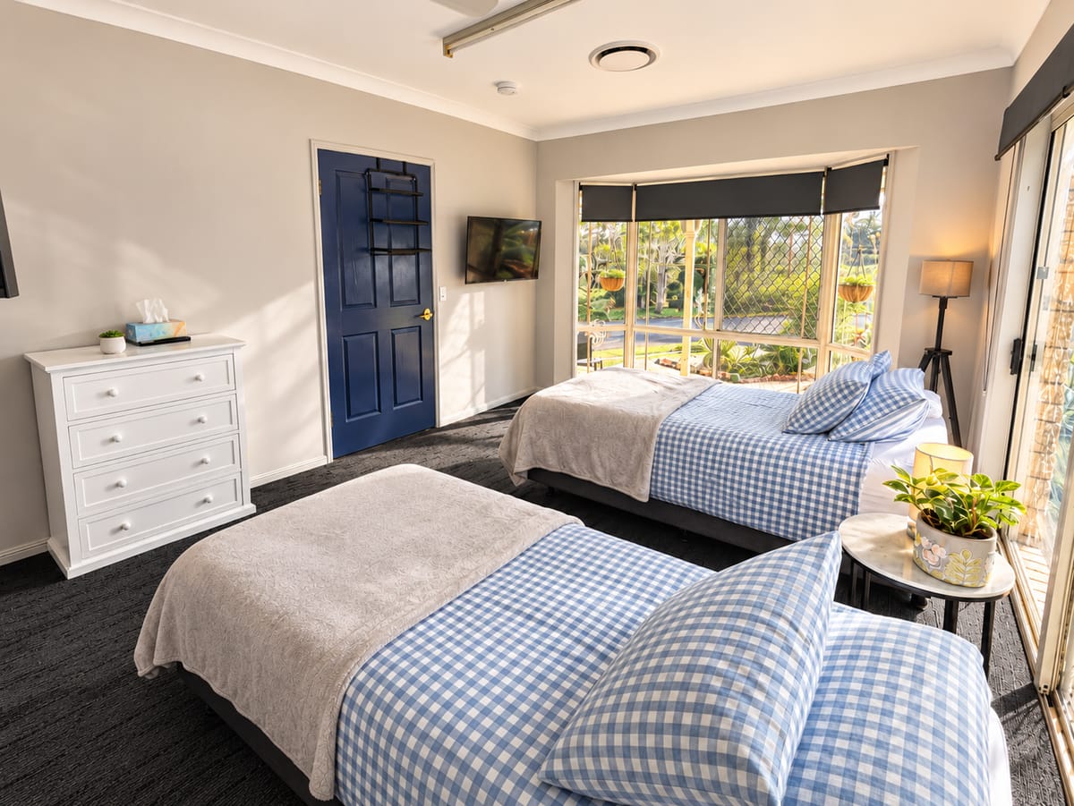 A room at our Alexandra Hills short stay house with a blue door, a television on the wall, a chest of drawers and two beds made in blue gingham.