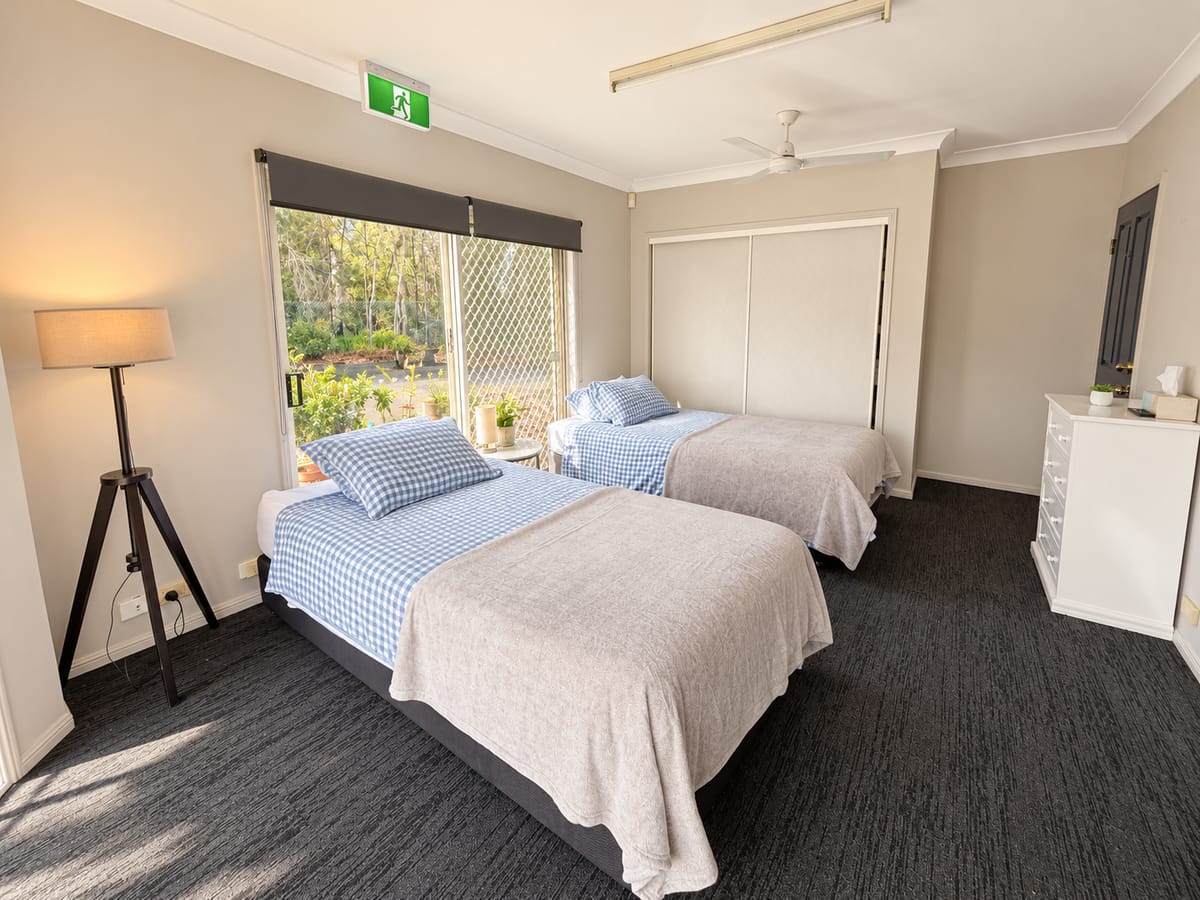 A twin room at our Alexandra Hills short stay house, with two single beds made in blue gingham, a floor lamp and a chest of drawers.
