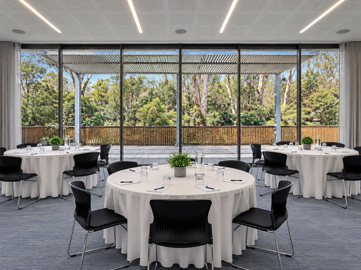 A function room with round tables in white linen and black chairs, and a glass wall looking out to a covered deck and bushland.